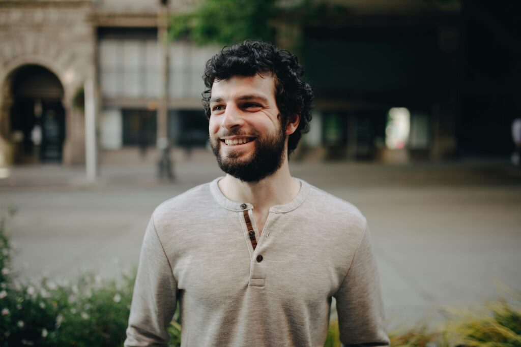 A bearded man with curly hair smiling outdoors in a casual setting.